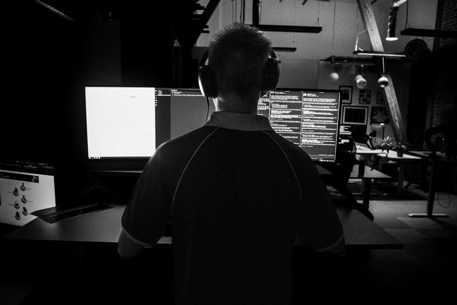 Black and white photo of Felix from behind, wearing headphones, working at three monitors in a dark loft office